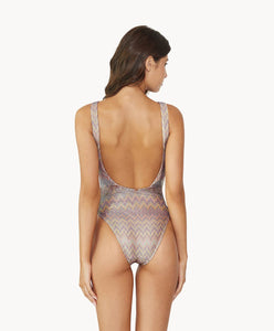 Brunette woman wearing a multi-colored print one piece swimsuit facing backwards towards a white wall.