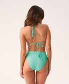 A woman wearing a teal triangle top bikini with matching high waist bottoms facing towards a white wall.