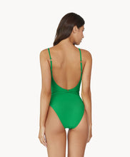 Brunette woman wearing green one piece swimsuit with gold beading facing backwards towards white wall.