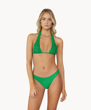 Blonde woman wearing a green halter bikini with net macramé & shimmery gold accents stands in front of white wall.