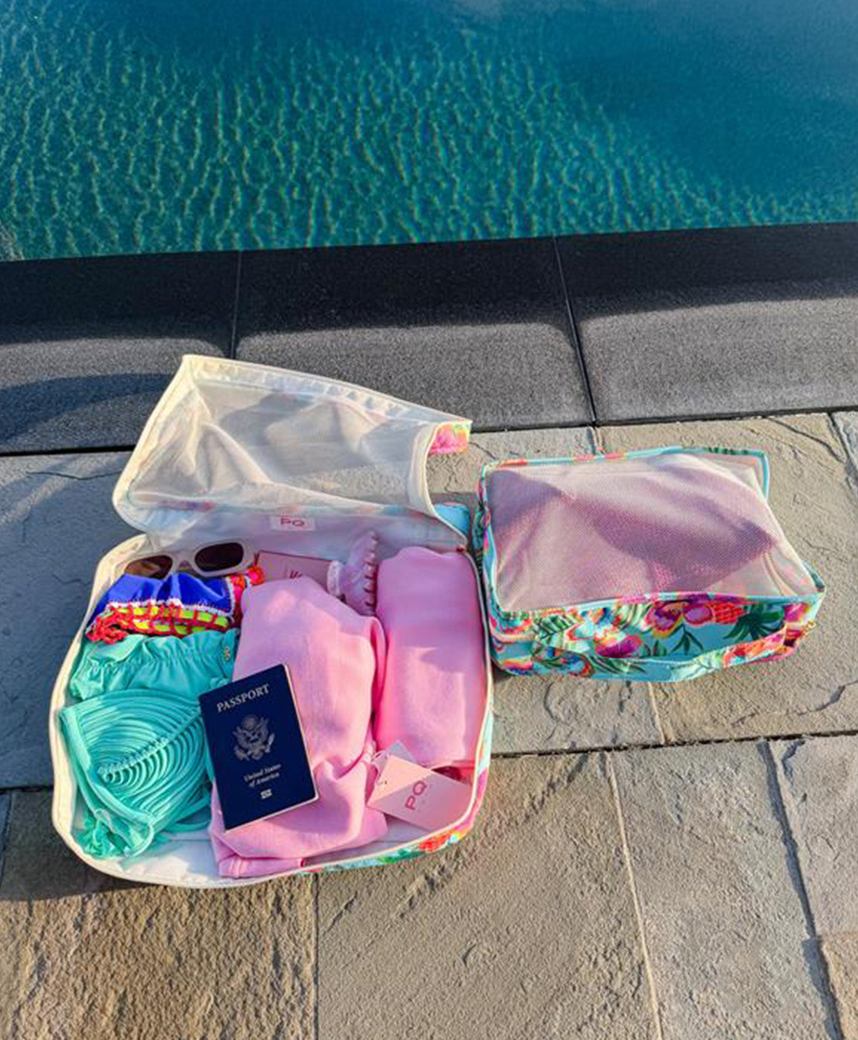 Open packing cube with travel items including a passport, sunglasses, and clothes by a pool.
