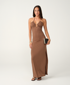 A woman modeling a floor-length brown halter-neck maxi dress with a side slit, standing against a white studio background and holding a small black clutch.