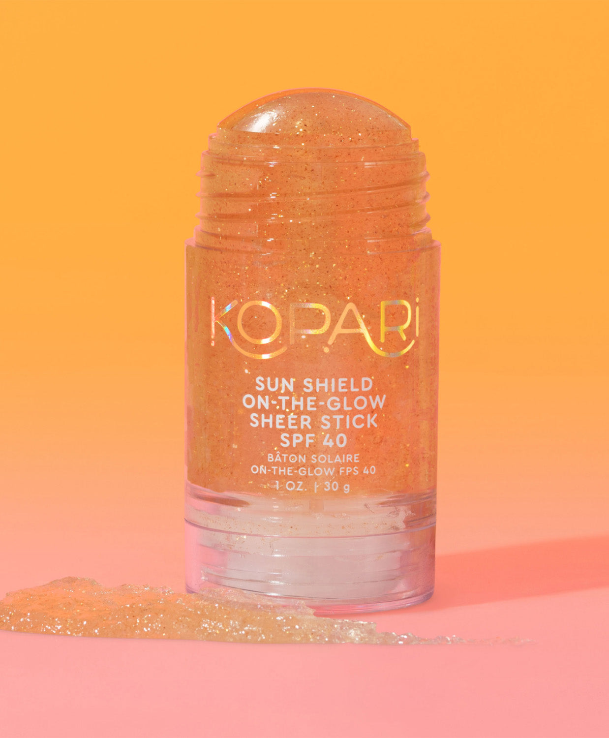 The Kopari Sun Shield On-the-Glow Sheer Stick SPF 40 standing upright with a swatch of product on an orange-pink background.
