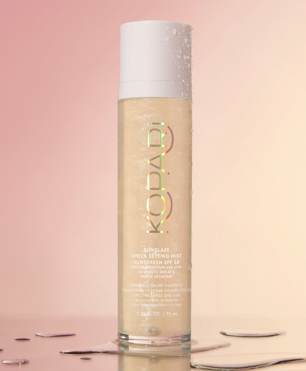 A close-up of the Kopari Sun Shield Body Glow SPF 50 spray bottle standing upright with water droplets on a pink background.