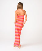 A woman in a long red, pink, and orange shimmery ombre strapless tube dress facing away in front of a white background.