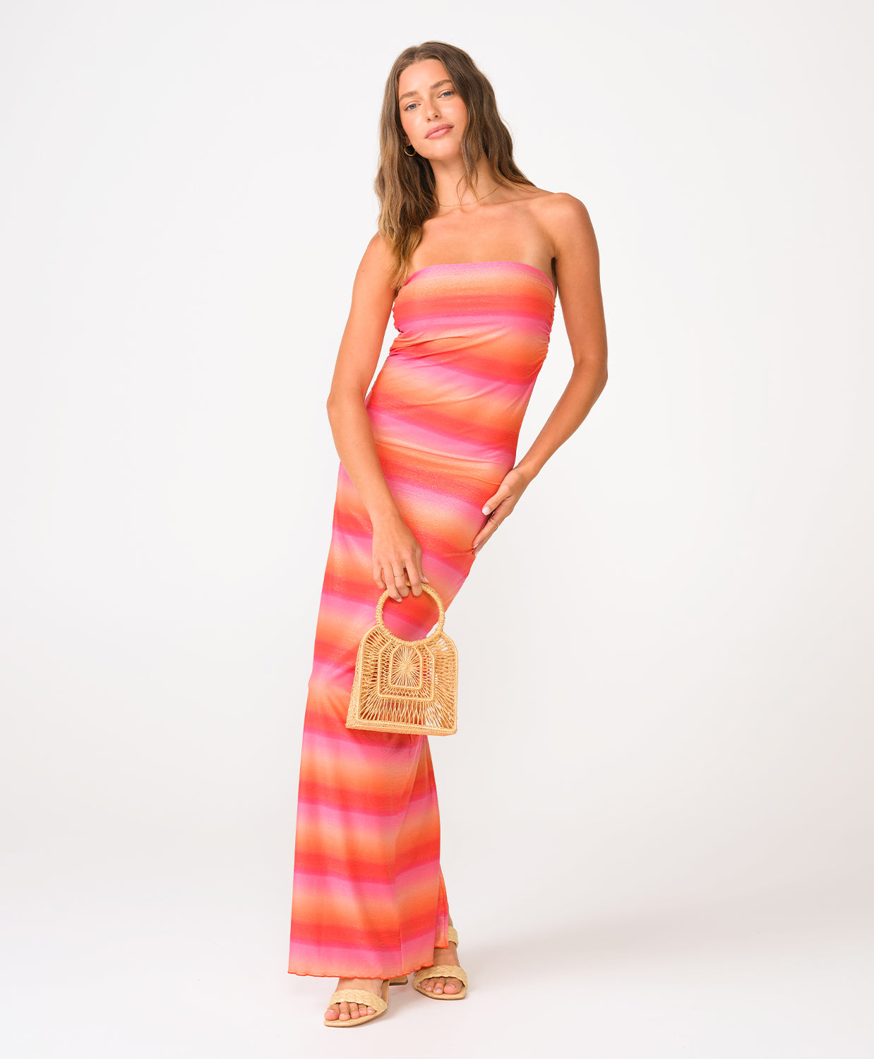 A woman wearing a red, pink, and orange shimmery strapless ombre tube dress holding a woven bag in front of a white background.