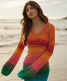 A woman wearing a long sleeve, knit, multi-colored dress standing on the beach near the ocean.