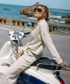 Woman in a beige outfit sitting on a scooter by the sea.