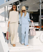 Two women walking on a boat, one in a beige outfit and the other in a light blue outfit, holding handbags.