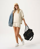 Woman holding a black bag, wearing a beige outfit with a denim jacket on a white background