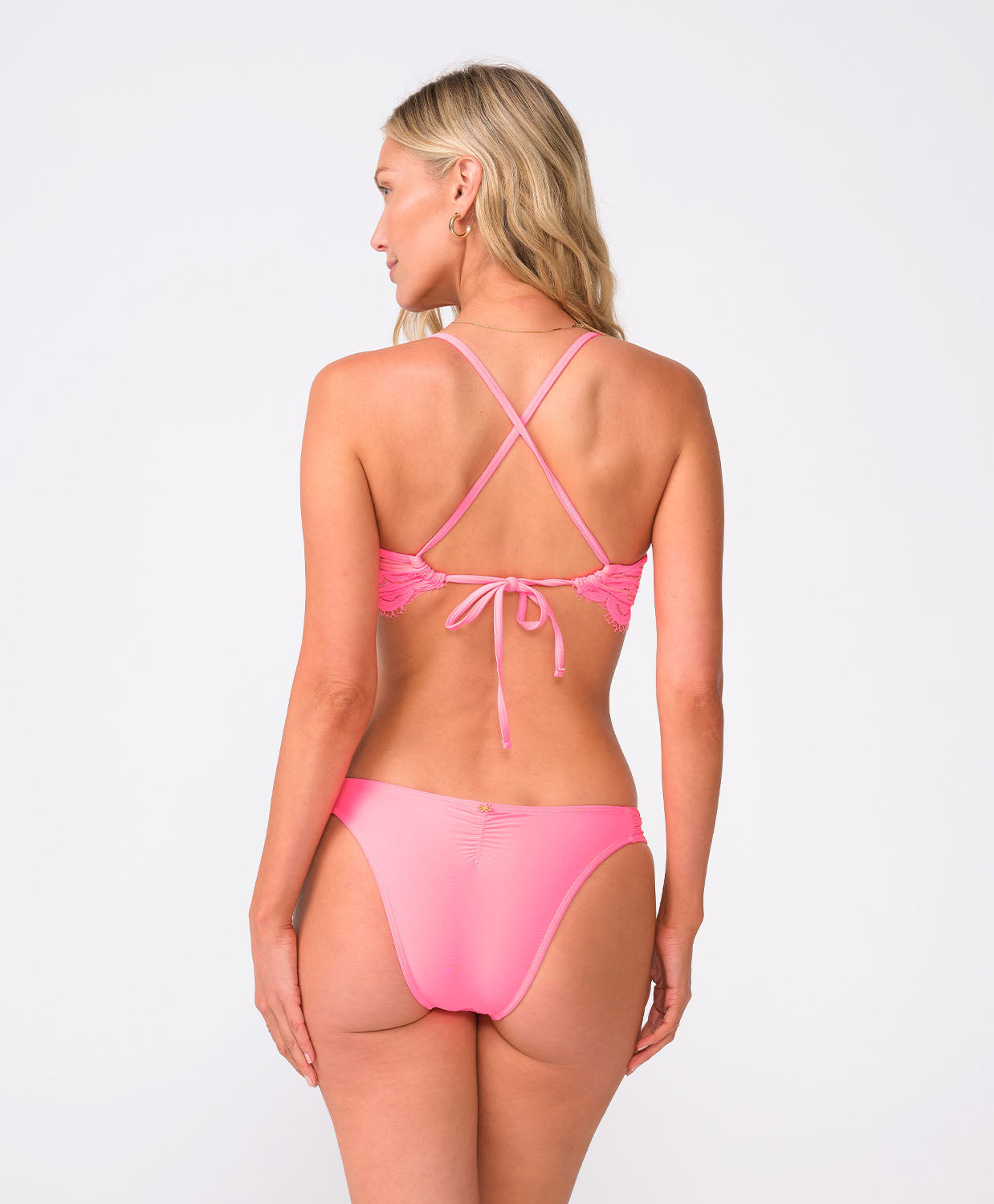 A woman wearing a pink lace bikini top and matching bottoms facing towards a white wall.