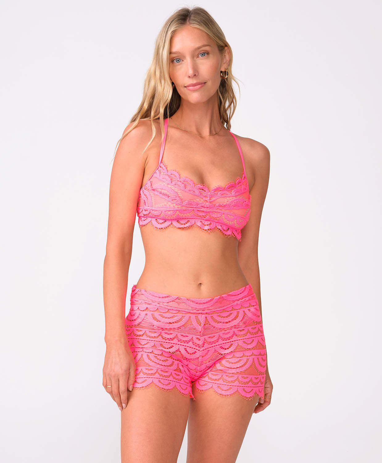 A woman wearing a pink lace bikini top and matching shorts standing against a white wall.
