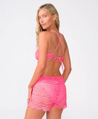 A woman wearing a pink lace bikini top and matching shorts standing against a white wall.