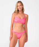 A woman wearing a pink lace bikini top and matching shorts standing against a white wall.