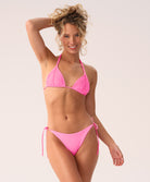 A woman wearing a pink triangle bikini top and tie-side bikini bottom standing against a white wall. 
