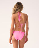 A woman wearing a pink triangle bikini top and tie-side bikini bottom facing towards a white wall. 