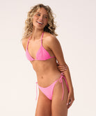 A woman wearing a pink triangle bikini top and tie-side bikini bottom standing against a white wall. 