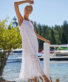 A blonde woman wearing a long white dress standing near a white fence and water. 