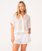 A woman wearing a matching white linen shirt and short set standing against a white wall.