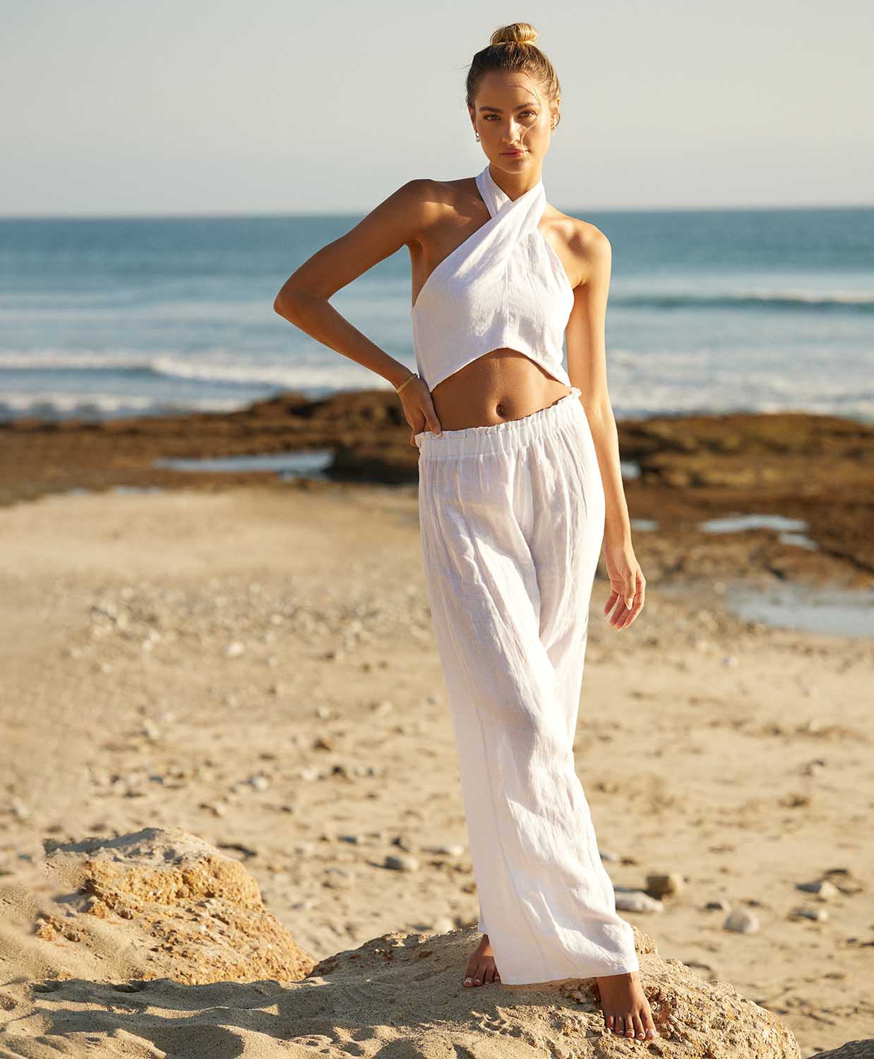 A woman wearing a white linen top and long linen pants standing on a beach near the ocean.