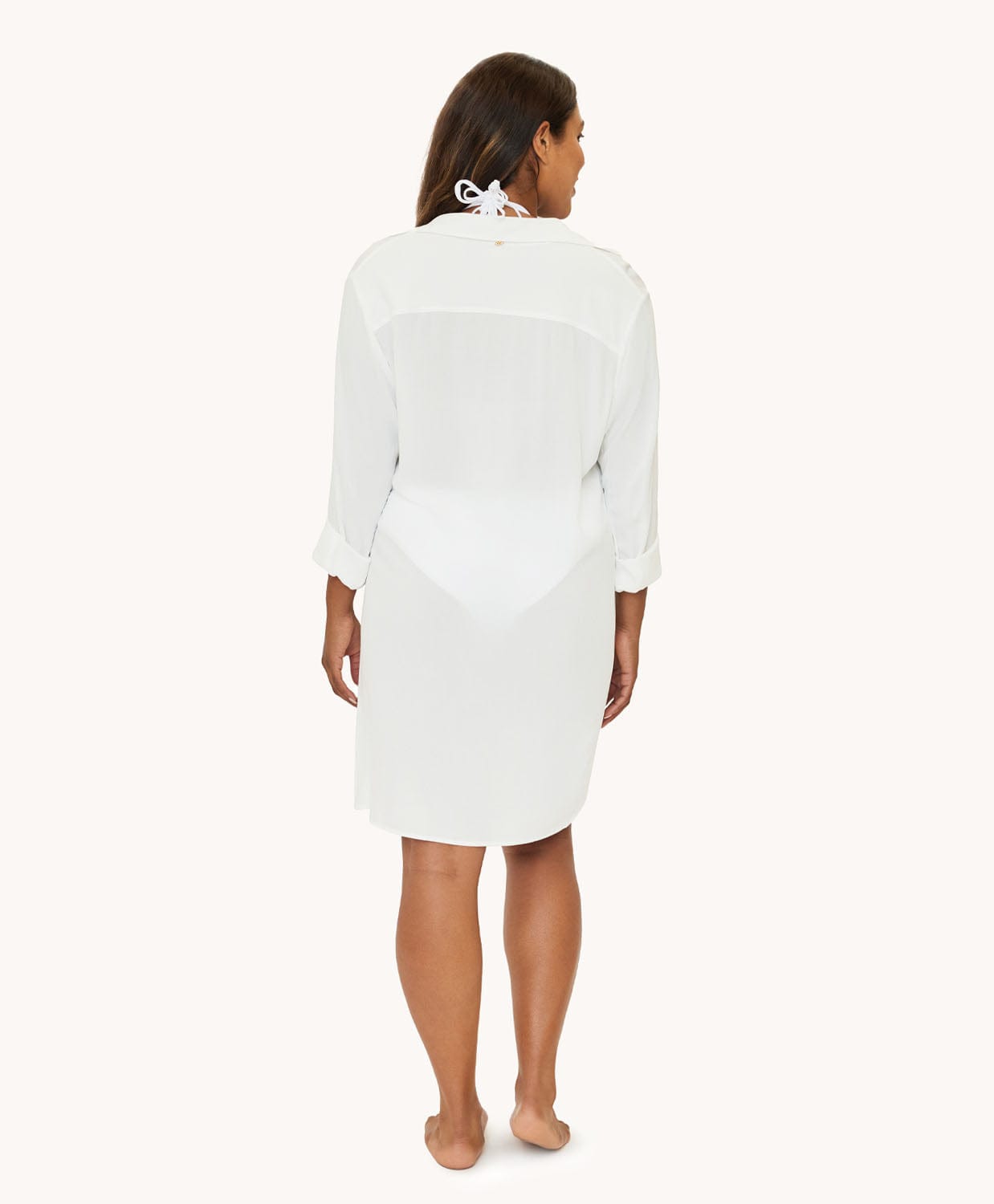 A woman wearing a white long sleeve cover up facing towards a white wall. 