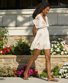 A woman wearing a short white dress with floral details walking on a sidewalk near flowers.