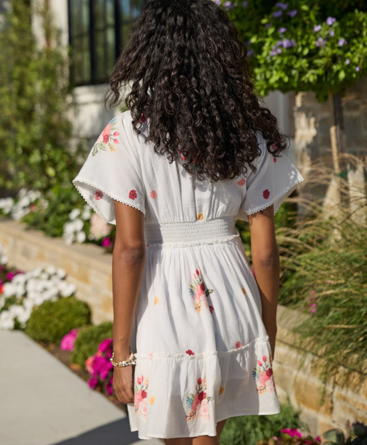 A woman wearing a short white dress with floral details walking on a sidewalk near flowers.