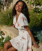 A woman wearing a short white dress with floral details sitting on a concrete ledge.