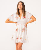 A woman wearing a short sleeve dress with floral details standing against a white wall.