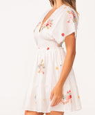 A woman wearing a short sleeve dress with floral details standing against a white wall.