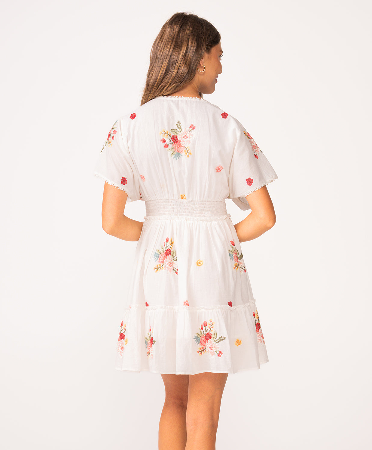 A woman wearing a short sleeve dress with floral details facing towards a white wall.