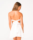A woman wearing a short white flowy dress with a tie in back facing away in front of a white background.