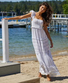 A brunette woman wearing a long white dress standing on the beach near water.