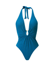 A textured turquoise halter one-piece swimsuit with gold details. Featured against a white wall background.