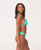 A woman wearing a turquoise triangle top bikini standing against a white wall.