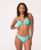 A woman wearing a turquoise triangle top bikini standing against a white wall.