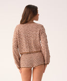 A woman wearing a long sleeve brown and beige striped top and matching shorts facing towards a white wall.