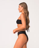 Woman wearing a black bandeau bikini top with gold detail and texture fabric in front of white wall.