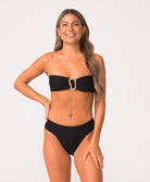 Woman wearing a black bandeau bikini top with gold detail and texture fabric in front of white wall.