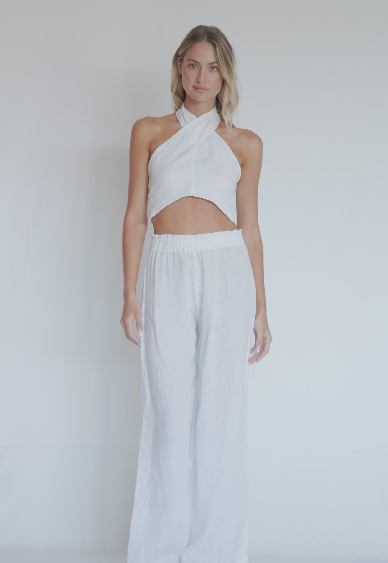 A woman wearing a white linen top and long linen pants spinning in front of a white wall.