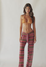 Brunette woman wearing multi-colored stripe print textured pants with a gold triangle bikini top spinning around in front of a white wall.