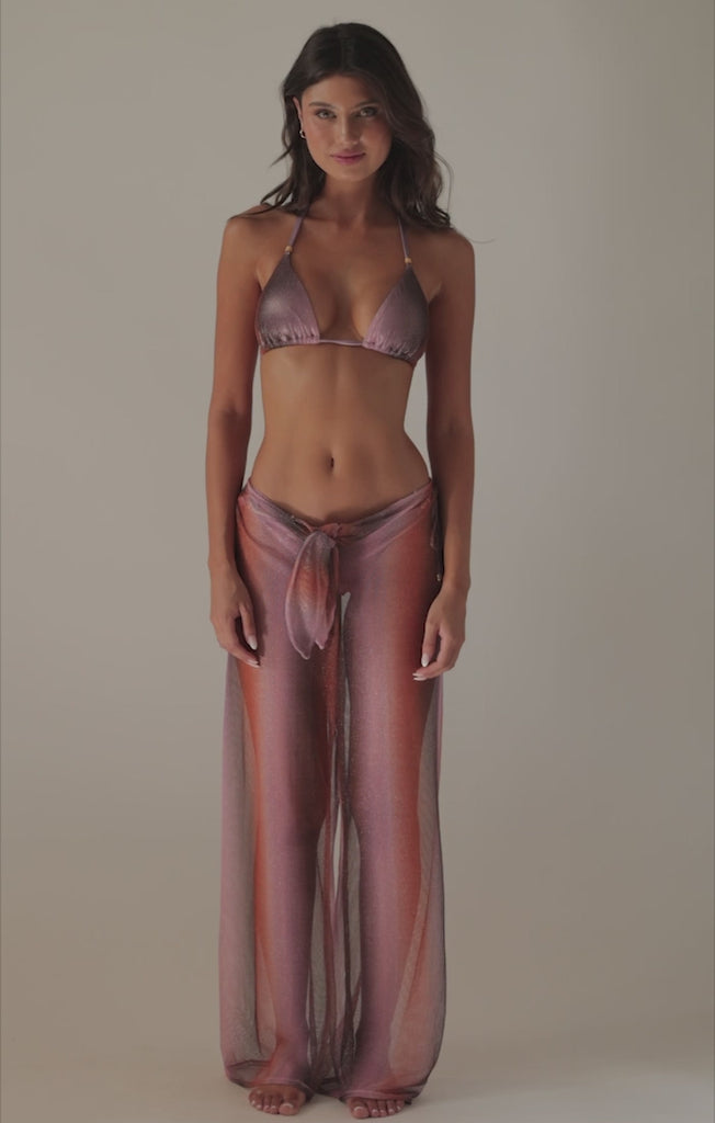 A brunette woman wearing a purple bikini and pants spinning in front of a white wall. 