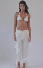A brunette woman wearing a white bikini top and white pants with a side slit spinning in front of a white wall. 