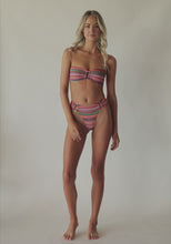 Blonde woman wearing a multi-colored stripe print bandeau bikini with gold details spinning around in front of a white wall.