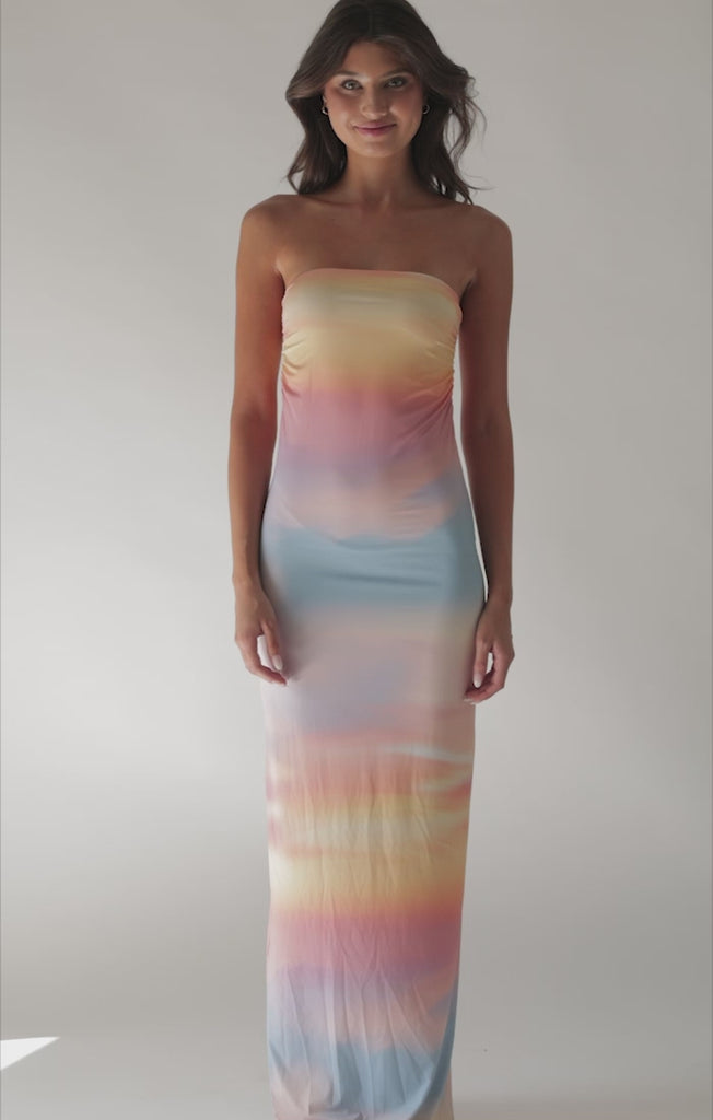 A brunette woman wearing a multi colored floor length dress spinning in front of a white wall. 