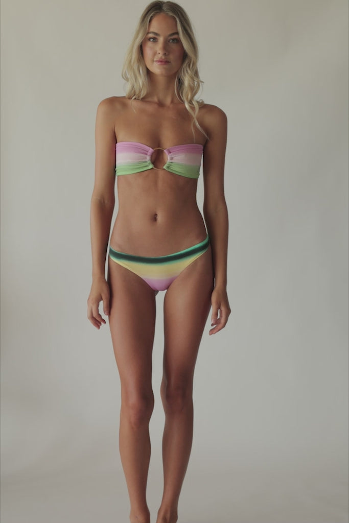 Blonde woman wearing a multi-colored stripe print bandeau bikini with gold details spinning around in front of a white wall.