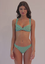 A brunette woman wearing a blue and green checkered bikini spinning in front of a white wall.