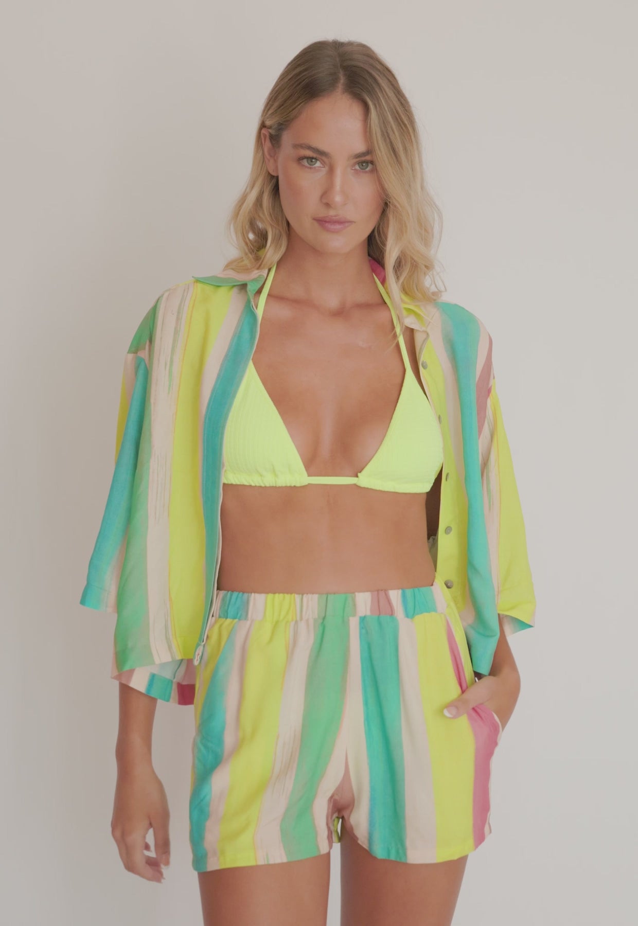 A woman wearing a green triangle top bikini and a colorful matching shirt and short set spinning in front of a white wall.