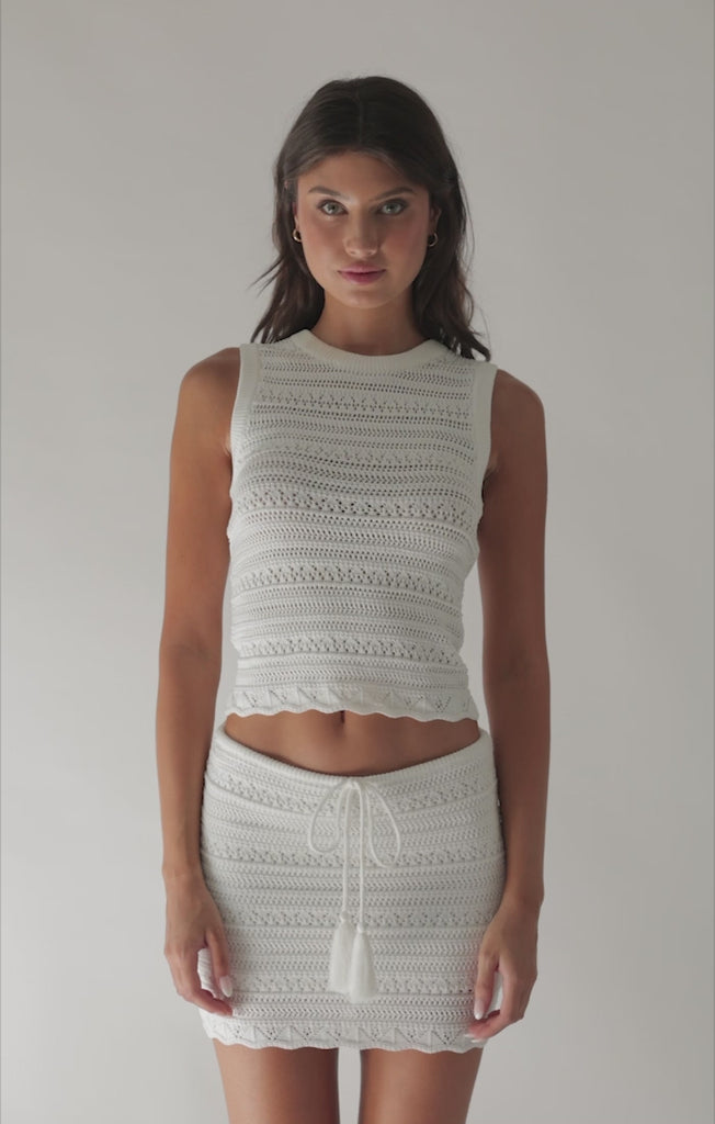 A brunette woman wearing a white crochet top and skirt spinning in front of a white wall. 