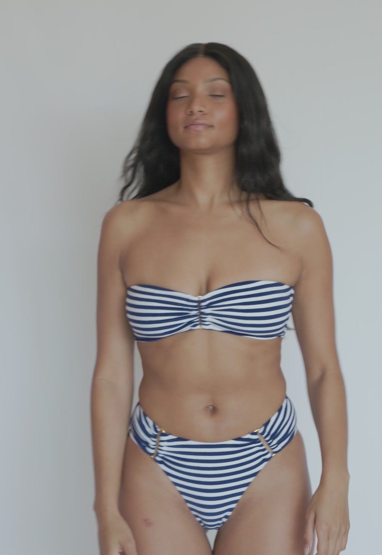 A woman wearing a blue and white striped bikini spinning in front of a white wall. 